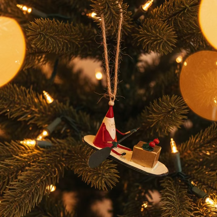 Decorative ornament of Santa Claus on a snowboard with a gift, hanging against a plain background.