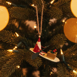 Decorative ornament of Santa Claus on a snowboard with a gift, hanging against a plain background.